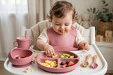 Pink silicone baby feeding set on high chair tray with bib, plate, cup and utensils